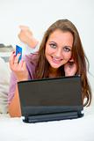 Smiling pretty woman lying on sofa and using credit card to shop from net
