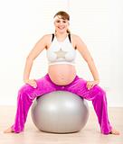 Smiling beautiful pregnant female doing pilates exercises on gray ball at home
