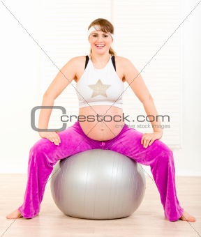 Smiling beautiful pregnant female doing pilates exercises on gray ball at home
