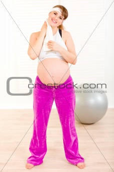 Smiling pregnant woman wiping her face with towel after exercising at home
