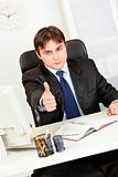 Confident modern businessman sitting at office desk and showing thumbs up gesture
