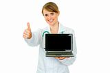 Smiling  medical doctor woman holding laptop with blank screen and showing thumbs up gesture
