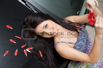 Young woman with flower lying back on black hood in petals of New car