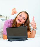 Smiling pretty woman lying on sofa with laptop and showing thumbs up gesture
