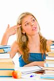 Stressful teen girl sitting at table with books and holding gun shaped hand near head
