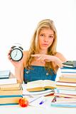 Worried teengirl sitting at table with books and pointing finger on alarm clock
