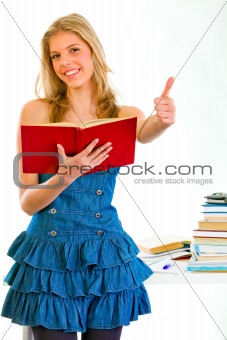 Smiling girl with book in hand standing near table and showing thumbs up gesture
