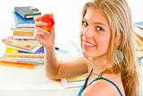 Smiling teen girl sitting at desk with books and holding apple in hand

