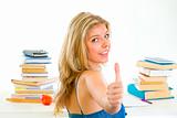 Smiling teen girl sitting at desk with books and showing thumbs up gesture
