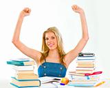 Pleased young girl with raised hands sitting at desk with books
