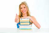 Smiling teen girl sitting at table with books  and showing thumbs up gesture
