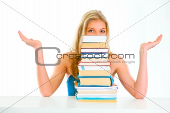 Sitting at table confused teen girl spreading hands apart and looking out of pile of book
