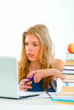 Pensive young girl sitting at table with books and looking on laptop

