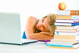 Tired young girl sleeping on table with books and laptop
