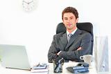 Smiling modern businessman with crossed arms on chest sitting at office desk
