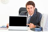 Smiling businessman sitting at office desk and pointing finger on laptop with blank screen
