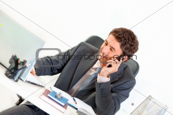 Sitting at office desk smiling businessman talking on mobile and looking on laptop
