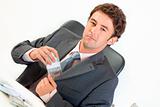 Confident businessman sitting at office desk and putting money in pocket
