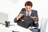 Shocked businessman sitting at office desk and looking in briefcase 
