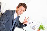 Smiling businessman sitting at office desk with laptop and showing victory gesture
