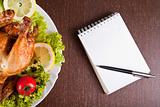 Restaurant table with roast chicken, notebook and pen 