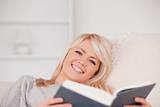 Smiling blonde woman reading a book while lying on a sofa