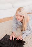 Pretty young blond woman relaxing on laptop lying on a carpet