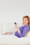 Smiling red-haired woman using a laptop and a phone while lying 