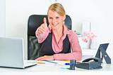 Smiling business woman sitting at office desk and  showing thumbs up gesture
