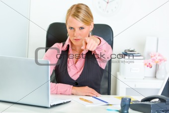 Confident business woman sitting at office desk and pointing finger at you
