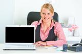Smiling business woman sitting at office desk and showing laptop with blank screen
