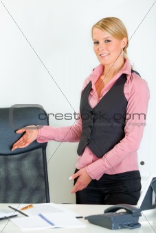 Smiling business woman standing near office desk and inviting to sit on chair
