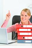 Sitting at office desk with pile of folders smiling business woman showing idea gesture
