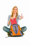 Sitting on floor smiling girl geting book from schoolbag and showing thumbs up gesture
