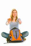 Smiling girl sitting on floor with schoolbag holding apple in hand and showing thumbs up gesture
