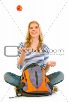 Smiling teengirl sitting on floor with schoolbag and throwing apple up
