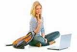 Pleased teengirl sitting on floor with schoolbag and laptop
