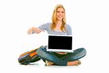 Smiling teengirl sitting on floor with schoolbag and pointing finger on laptop with blank screen
