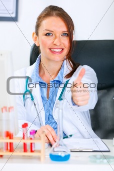 Smiling doctor woman sitting at office table and showing thumbs up gesture
