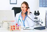 Smiling doctor woman sitting at office table and talking on phone

