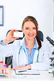 Smiling doctor woman sitting at office table and holding blank business card
