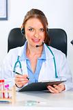 Smiling doctor woman with headset sitting at office table and holding  clipboard
