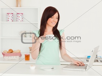 Smiling red-haired female relaxing with her laptop in the kitche
