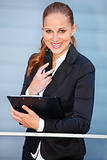 Leaning on railing at office building  smiling business woman holding mobile and clipboard 
