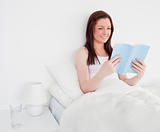 Pretty red-haired woman reading a book while sitting on her bed