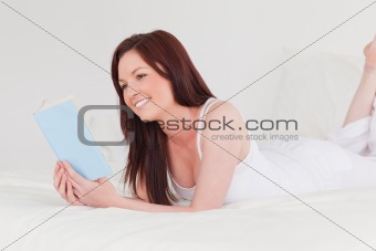 Gorgeous red-haired woman reading a book while lying on her bed