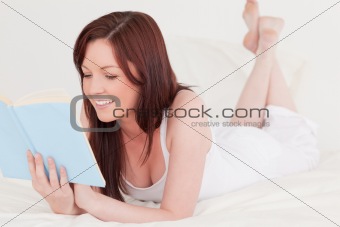 Cute red-haired woman reading a book while lying on her bed