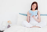Cute red-haired woman reading a book while sitting on her bed