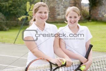 Two young girl friends with rackets on tennis court smiling