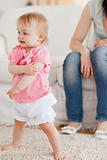 Baby standing on a carpet while her mother is sitting on a sofa
